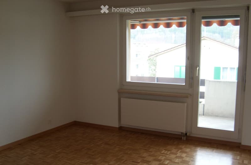 Empty room with wooden flooring, white walls, a window with blinds, and a radiator below it. A sliding glass door leads to a balcony or outdoor area.