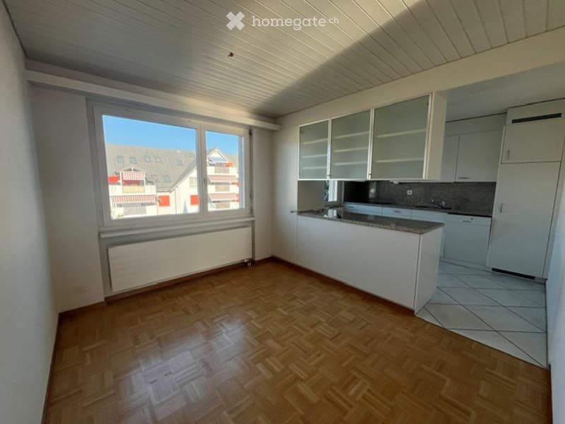 Empty kitchen with white cabinets, wooden flooring, and large windows overlooking a street.