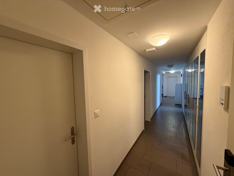 Clean, modern hallway with white walls, tiled floor, and recessed lighting. Features multiple doors and a glass-paneled wall on one side.