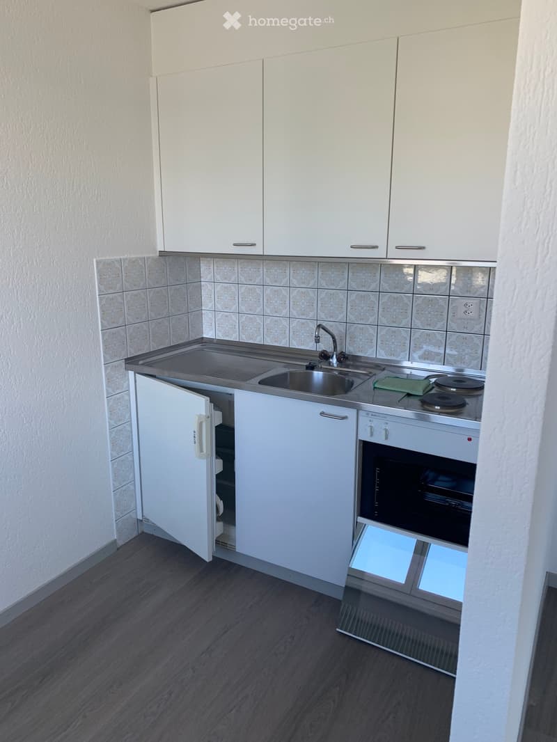 Modern kitchen with white cabinets, stainless steel sink, oven, and refrigerator. The walls are tiled with a floral pattern, and the floor is dark wood.