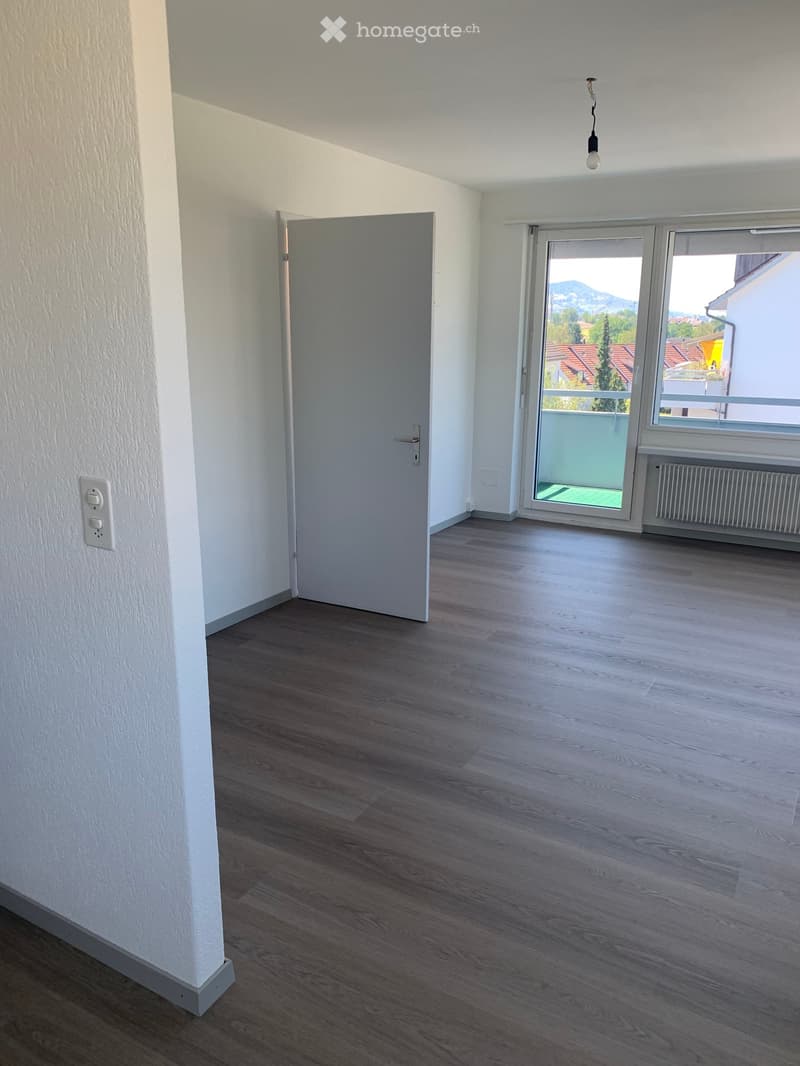 Empty room with wooden flooring, white walls, a closed door, and a large window with a view of the outside area.