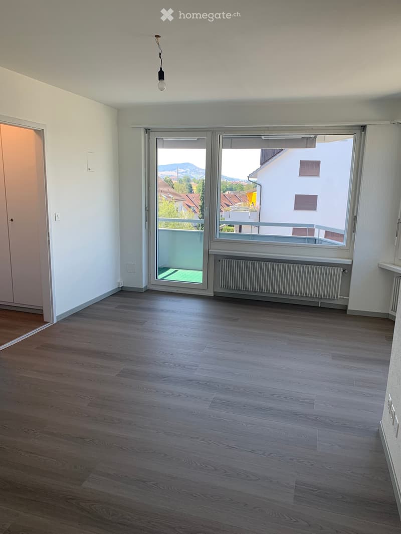 Empty room with wooden flooring, white walls, large windows, and a balcony access door.