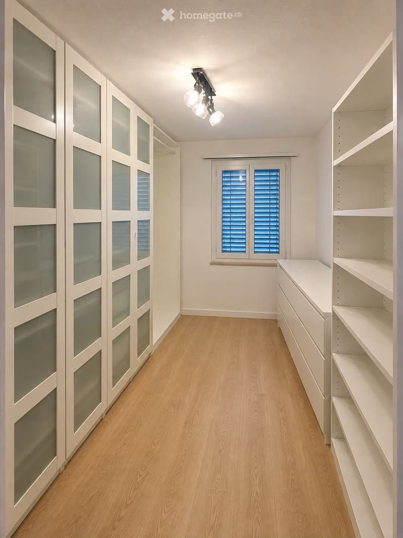 Empty room with light wooden flooring, white walls, built-in closet with mirrored doors, open shelving unit, and a window with blue shutters.