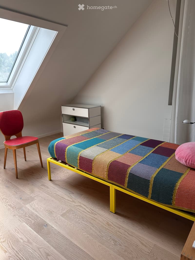 Modern bedroom with a colorful checkered bedspread, a wooden chair, and a sleek nightstand. The room features wooden flooring and large windows with a view.