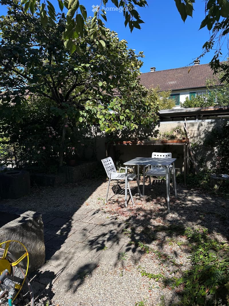 A small outdoor seating area with a table and two chairs, surrounded by greenery and a house in the background.