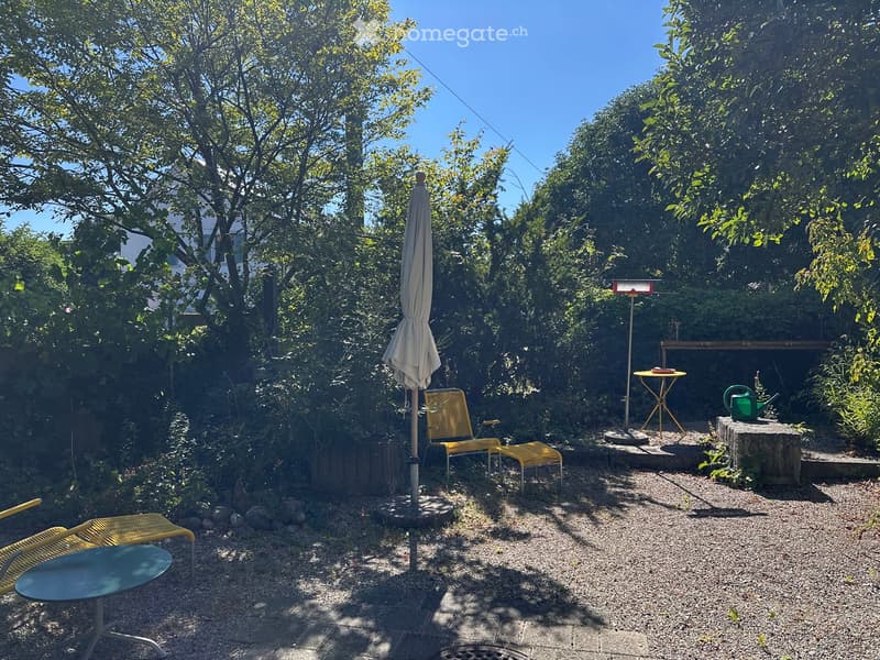 A small, well-maintained garden with gravel ground, surrounded by lush greenery. Features include a yellow lounge chair, a small round table, a closed umbrella, and a green watering can on a stone platform.
