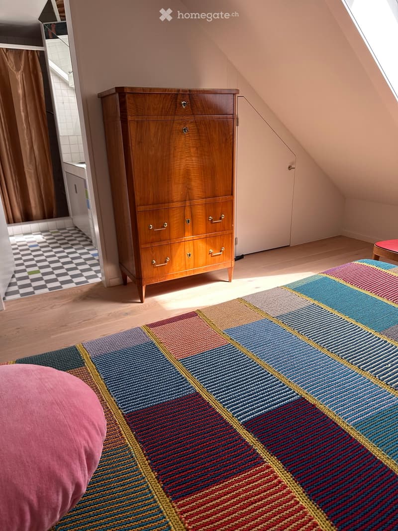 A cozy attic bedroom with wooden floors, a large wooden dresser, a colorful geometric rug, and a pink round cushion on the floor.