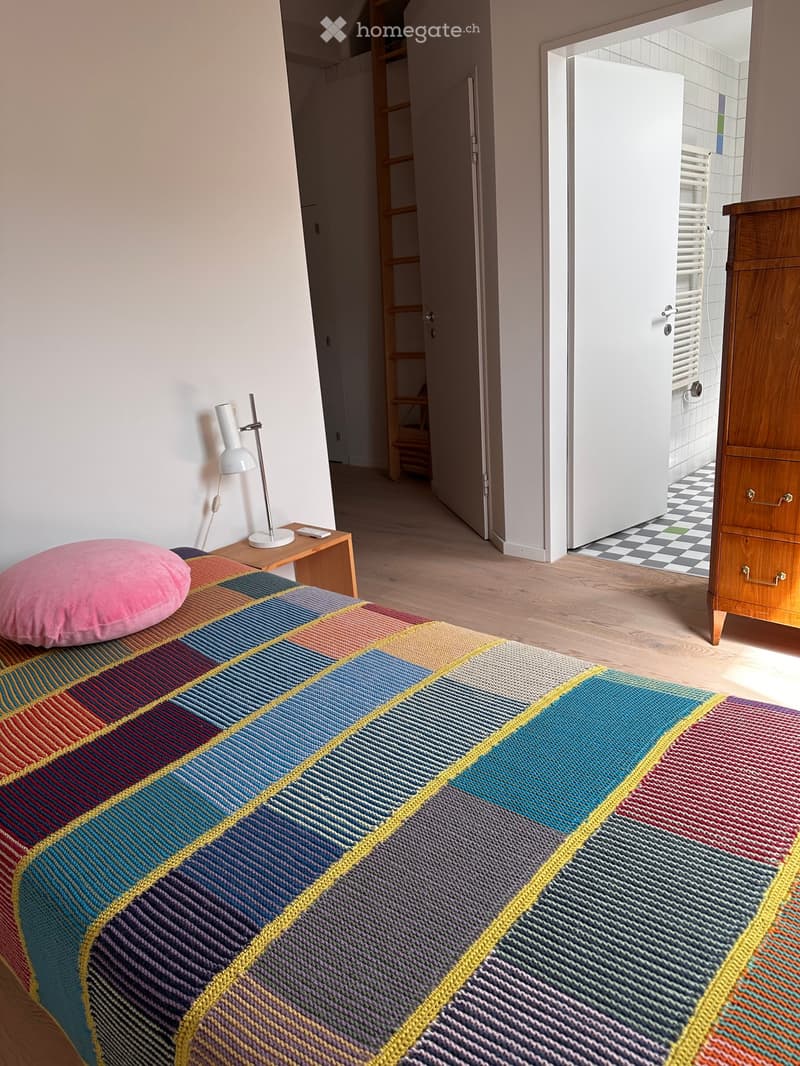 A modern bedroom with a colorful, patterned bedspread, a pink pillow, a wooden nightstand with a lamp, and an open door leading to a bathroom.