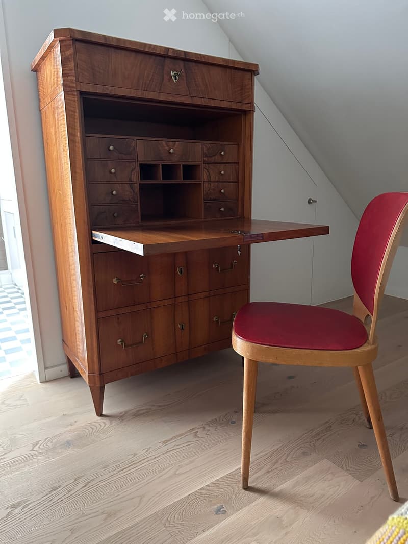 A wooden desk with multiple drawers and a pull-out writing surface, paired with a red upholstered chair, set against a slanted ceiling in a room with wooden flooring.