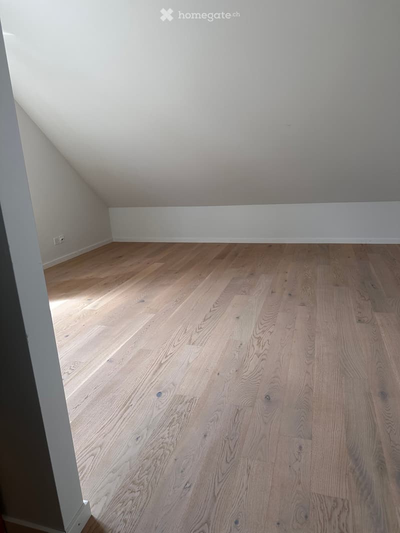 Empty room with light wooden flooring and white walls, featuring a slanted ceiling typical of an attic space.
