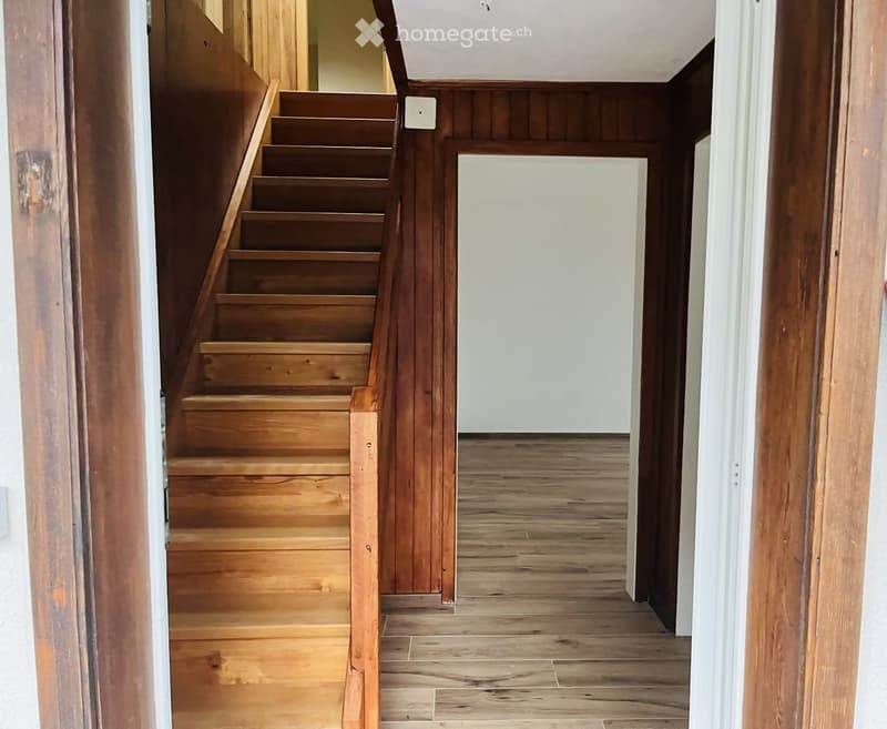 Wooden staircase leading upwards, wooden floor, white walls, open doorway to another room.