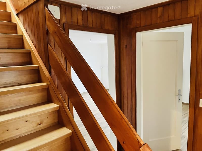 Wooden staircase with a handrail leading to an open doorway, with wooden panel walls and a light-colored floor visible in the background.