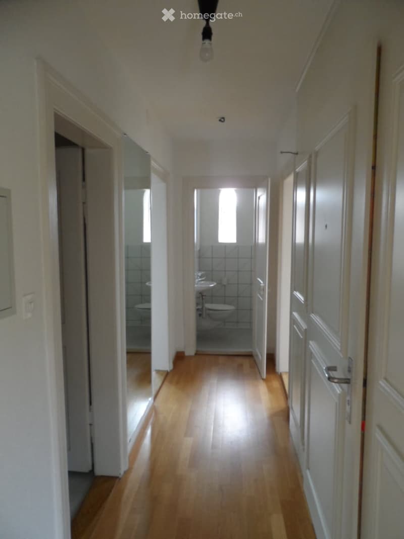 Empty hallway with wooden flooring, white walls, and several doors leading to rooms, including a visible bathroom.