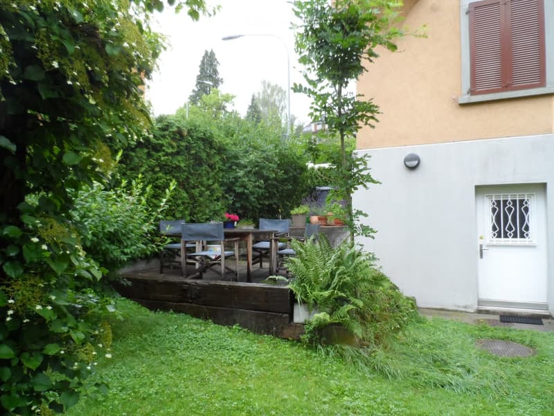 A small, well-maintained garden with a wooden deck, a table and chairs, potted plants, and a white door leading to the house.