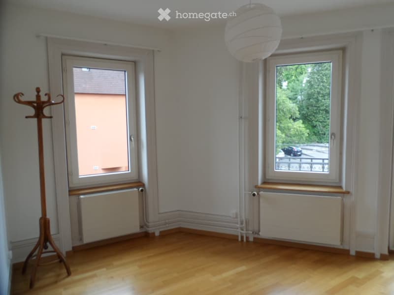 Empty room with wooden flooring, white walls, two windows, radiator, and a coat rack.