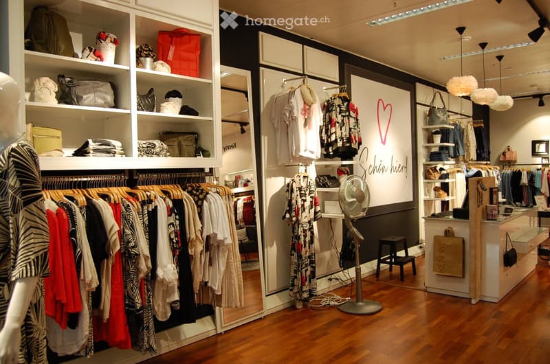 A stylish clothing store interior with wooden flooring, white shelving units filled with folded clothes and bags, clothing racks with various garments, a standing fan, and a small stool near the display area.