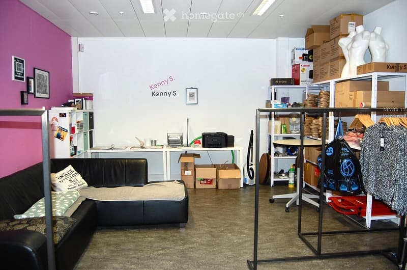 A furnished room with a black leather sofa, a pet bed, a table with office supplies, shelves with various items, mannequins, a clothing rack, and framed pictures on the walls.