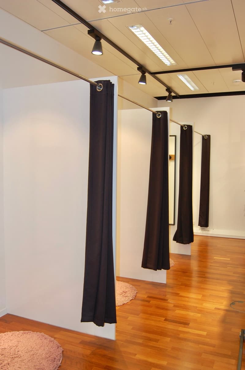 Empty room with wooden flooring, white walls, and dark curtains hanging from a track system. Minimalist design with recessed lighting and a small pink rug in the foreground.