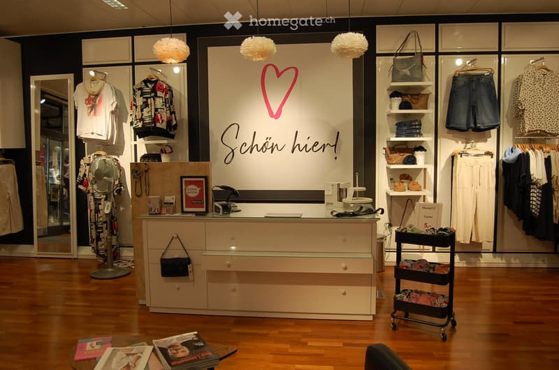 Retail clothing store interior with wooden flooring, clothing racks, shelves, a mirror, a fan, a table with magazines, and a cart with items.