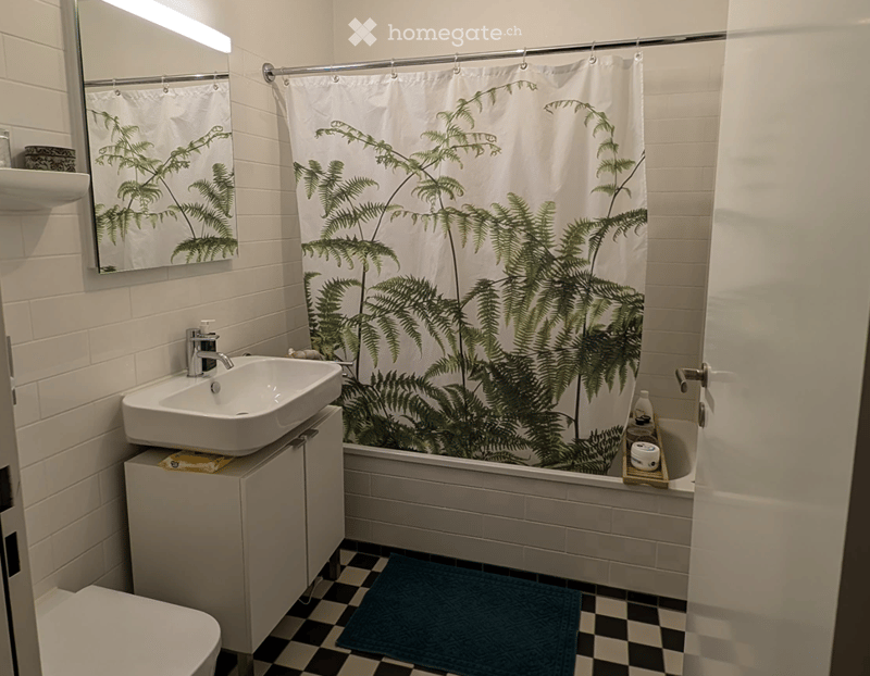 Modern bathroom with white tiles, a white sink with a chrome faucet, a bathtub with a green fern-patterned shower curtain, a mirror, a small cabinet, and a blue bathroom rug on a black and white checkered floor.