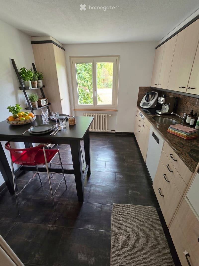 Modern kitchen with dark tiled floor, light wooden cabinets, and a black dining table with a red chair. The counter features a sink, coffee maker, and blender, with a bowl of fruit and cups on the table.