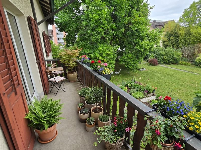 Balcony with wooden railing, potted plants, small table and chairs, overlooking a green garden.