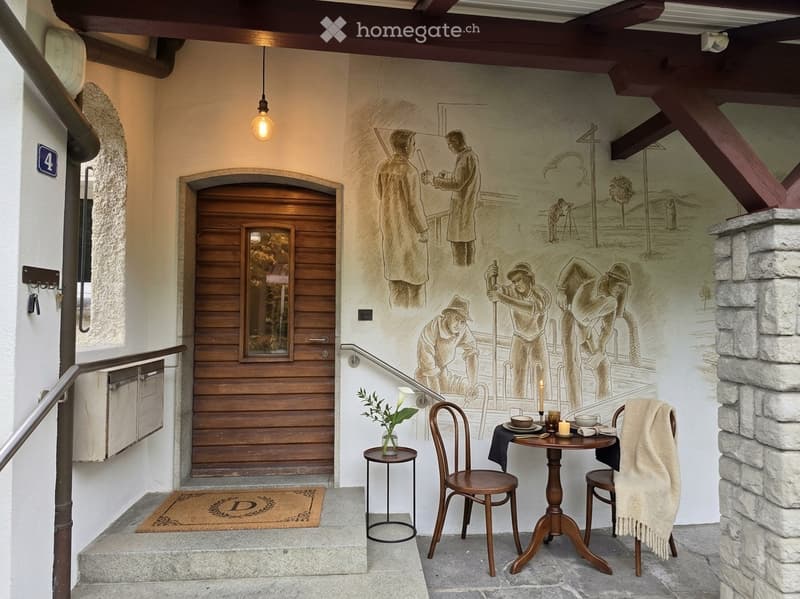 A cozy, furnished area with a small table, chairs, a vase with flowers, and a doormat. The wall features a large, detailed mural depicting historical figures and scenes.