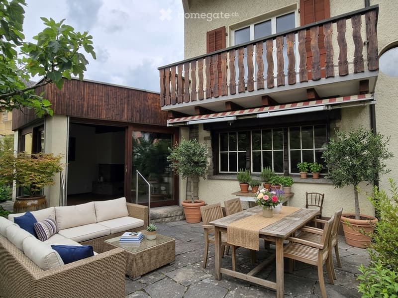 Outdoor patio with a wooden dining table, chairs, a wicker sofa, and potted plants. The area is adjacent to a house with a balcony and large windows.