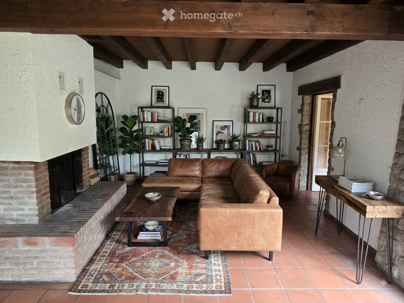 Living room with exposed wooden beams, brick fireplace, leather sofa, wooden coffee table, bookshelf, and decorative plants.