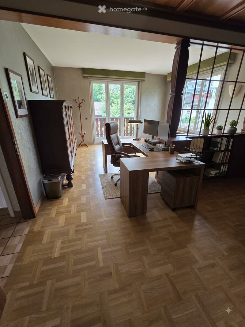 Spacious, well-lit office with wooden herringbone flooring, large windows, and a balcony access. Features a large wooden desk with a computer, office chair, lamp, and various office supplies. A tall wooden cabinet and bookshelf are present, along with a c
