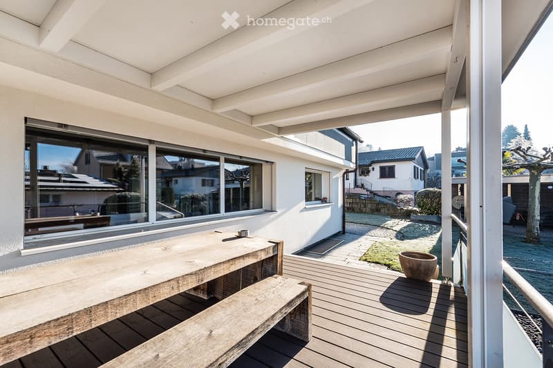 Wooden deck with a long table and bench, large windows overlooking a garden area with a small tree and potted plant.