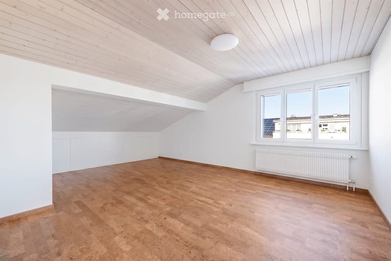 Empty room with wooden flooring, white walls, and a slanted wooden ceiling. Features include a large window with a view, a built-in cabinet, and a radiator.