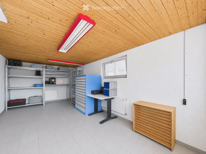 A well-lit, organized room with wooden slatted ceiling, tiled floor, and white walls. Features include metal shelving units, a standing desk, a wooden radiator, and various storage boxes.
