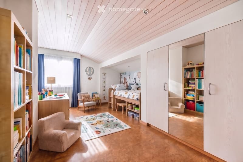 Bright, modern bedroom with wooden floors, slanted ceiling, built-in wardrobe, bed with colorful bedding, desk, and seating area.