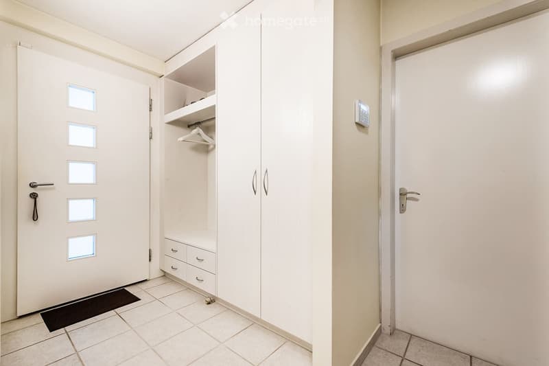 A clean, unfurnished hallway with tiled flooring, white walls, and a built-in closet with shelves and drawers. A door with frosted glass panels is visible on the left, and a small black doormat is placed in front of it.