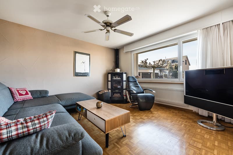 Modern living room with wooden flooring, large windows, a grey sectional sofa, a wooden coffee table, an armchair, a television, and a fireplace.