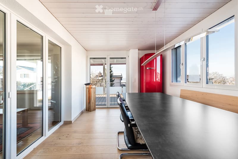 Modern, minimalist room with light wooden flooring, large windows, and a black dining table with chairs. A red refrigerator is visible, and there is a sliding glass door leading to a balcony area.