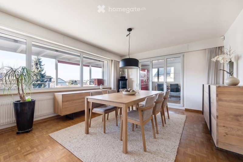 Bright dining area with a wooden dining table, six chairs, pendant light, and large windows overlooking a garden. Includes a tall plant in a pot and a decorative vase on a sideboard.