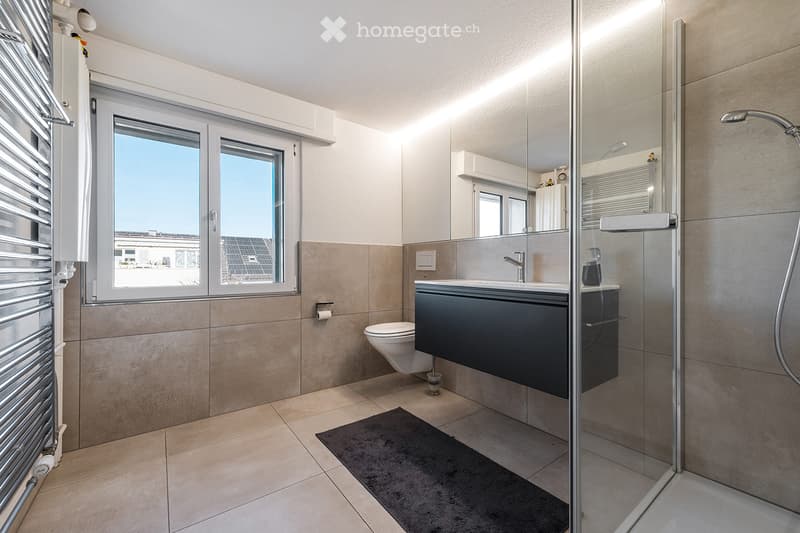 Modern bathroom with a toilet, sink, shower, and a large mirror. The space features tiled walls and floor, a radiator, and a window offering a view of the outside.