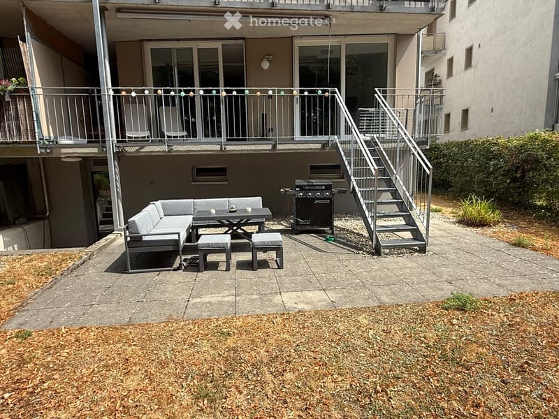 Outdoor patio with a grey sectional sofa, small table, two stools, and a grill. Metal staircase leads to an upper balcony with chairs and a railing.