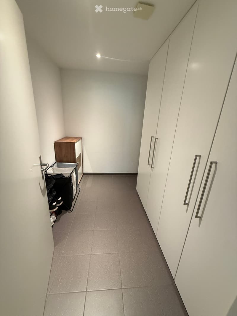 Narrow hallway with tiled floor, white walls, and built-in storage units on one side. A small wooden shelf and a shoe rack are visible on the left side.