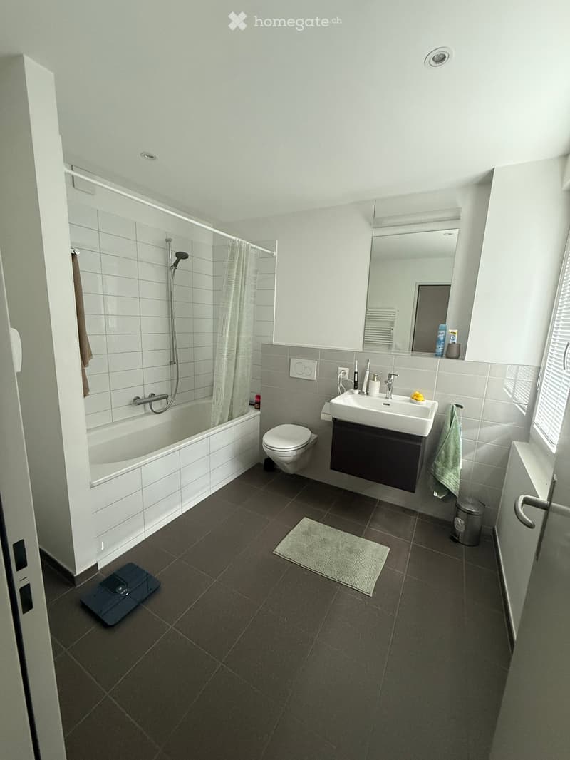 Modern bathroom with white tiles, bathtub, toilet, sink, mirror, towel, bathrobe, scale, and trash can.