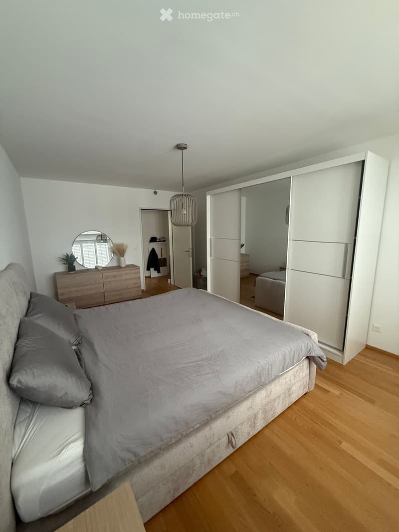 Modern bedroom with a large bed, grey bedding, a wooden dresser with a mirror, a wardrobe with mirrored doors, and a hanging pendant light.