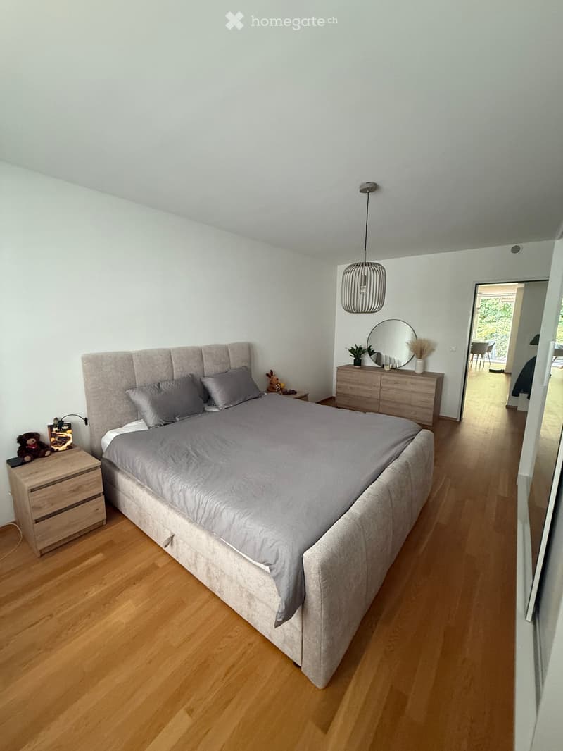 Modern bedroom with a large bed, grey bedding, wooden nightstand, dresser, mirror, and pendant light. The room features light wooden flooring and white walls.