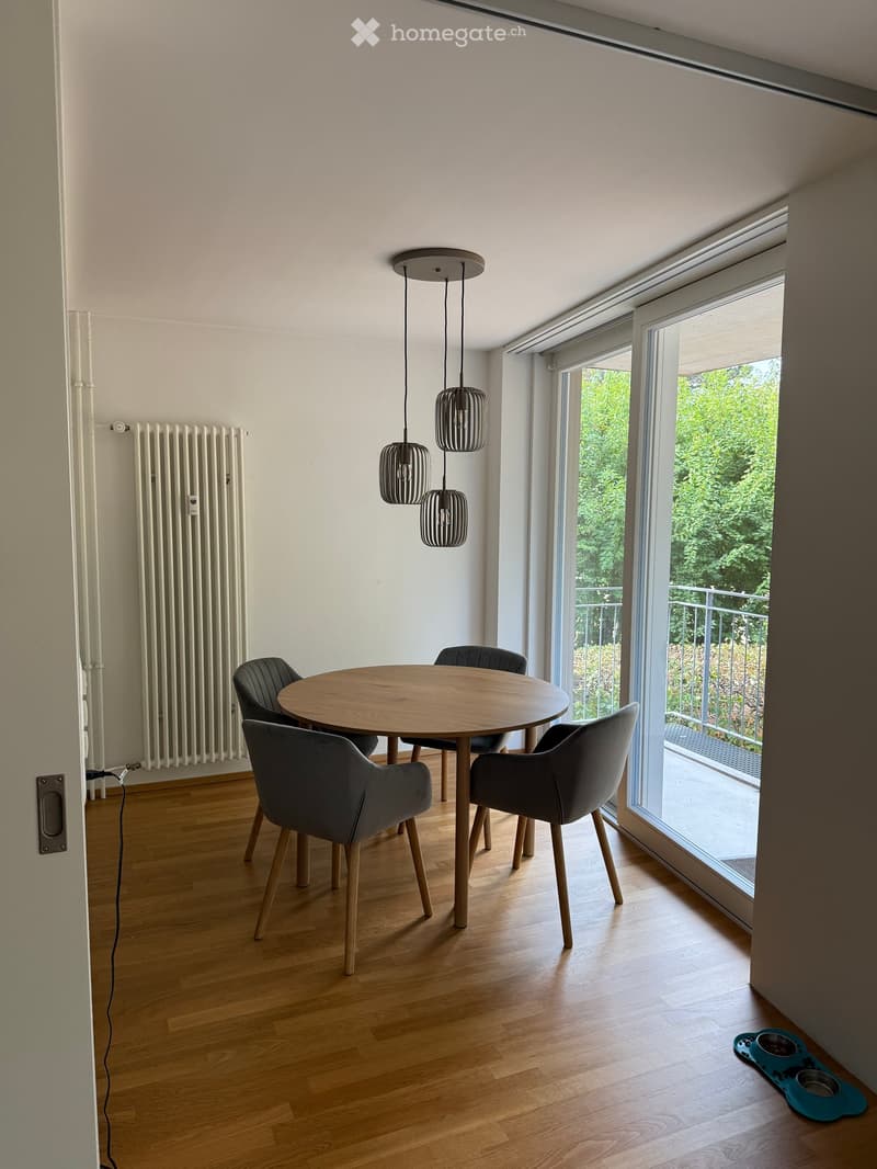 Dining area with a round wooden table, four grey chairs, pendant lights, and large windows overlooking a balcony.