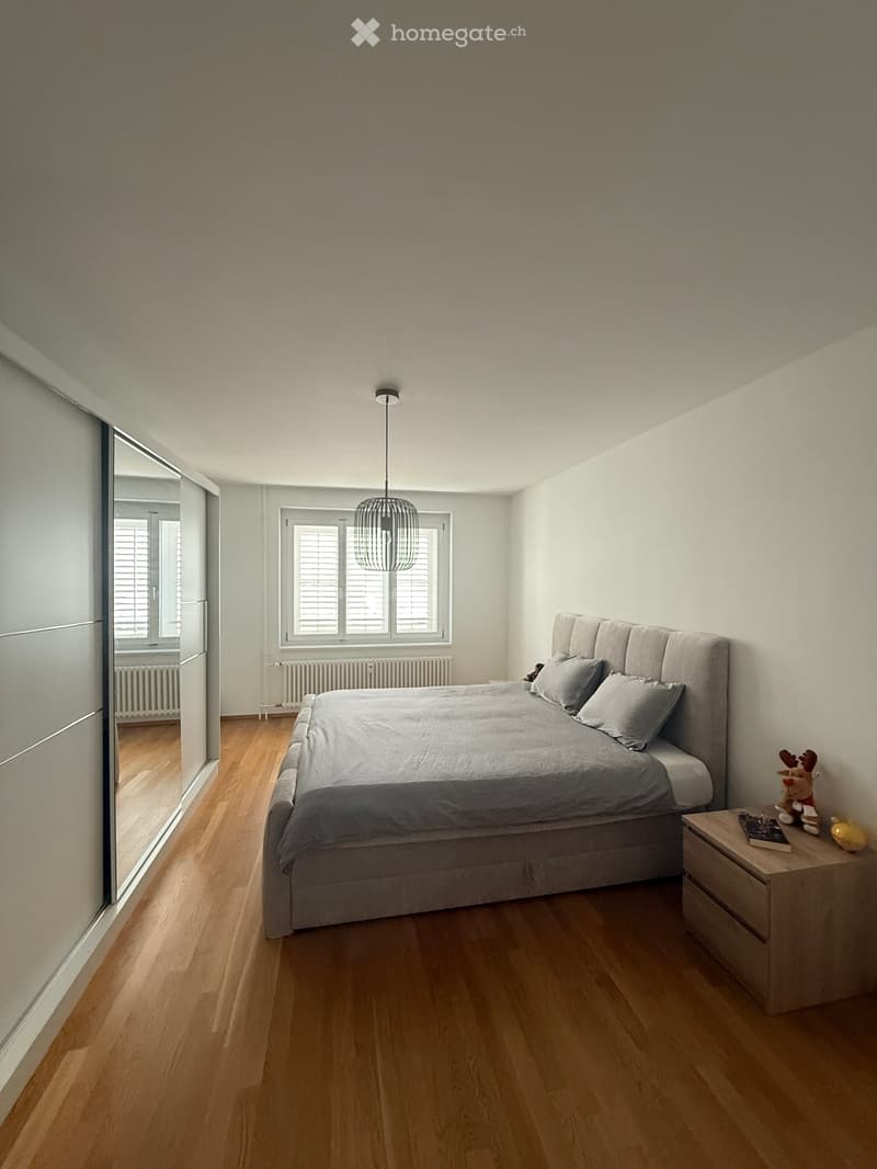 Modern bedroom with wooden flooring, large bed with grey bedding, nightstand with a toy and book, large window with blinds, and a hanging pendant light.