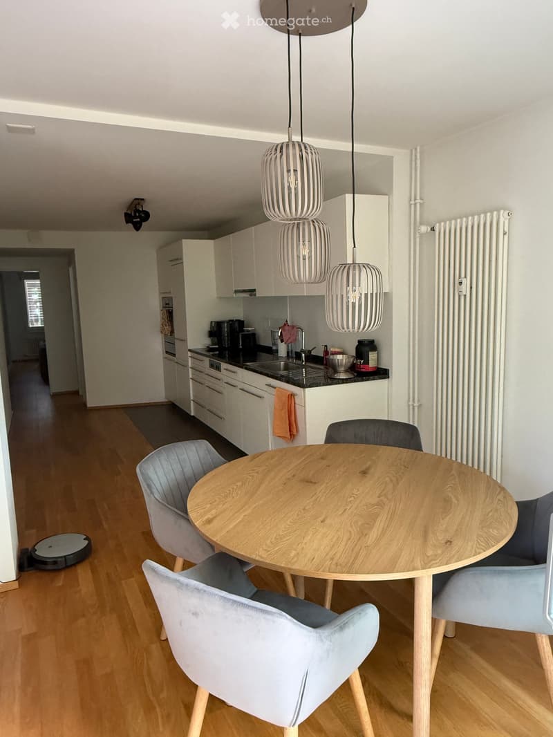Modern kitchen and dining area with light wood flooring, white cabinets, a round wooden dining table with four grey upholstered chairs, and pendant lights above the table. The kitchen features a black countertop with a sink, coffee maker, and other small