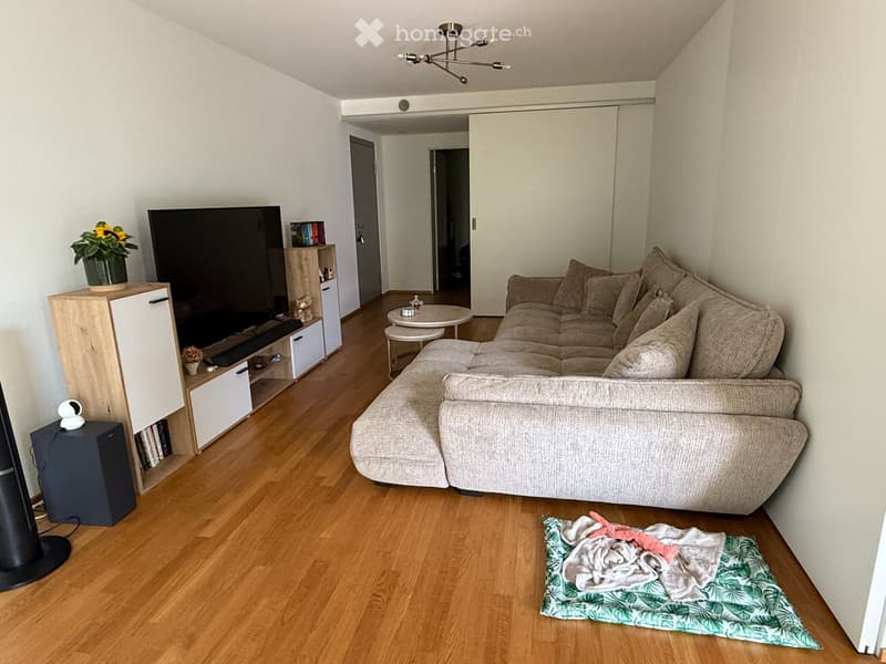 Modern living room with a sectional sofa, coffee table, TV stand, bookshelf, and a small round table. The room features wooden flooring and white walls.