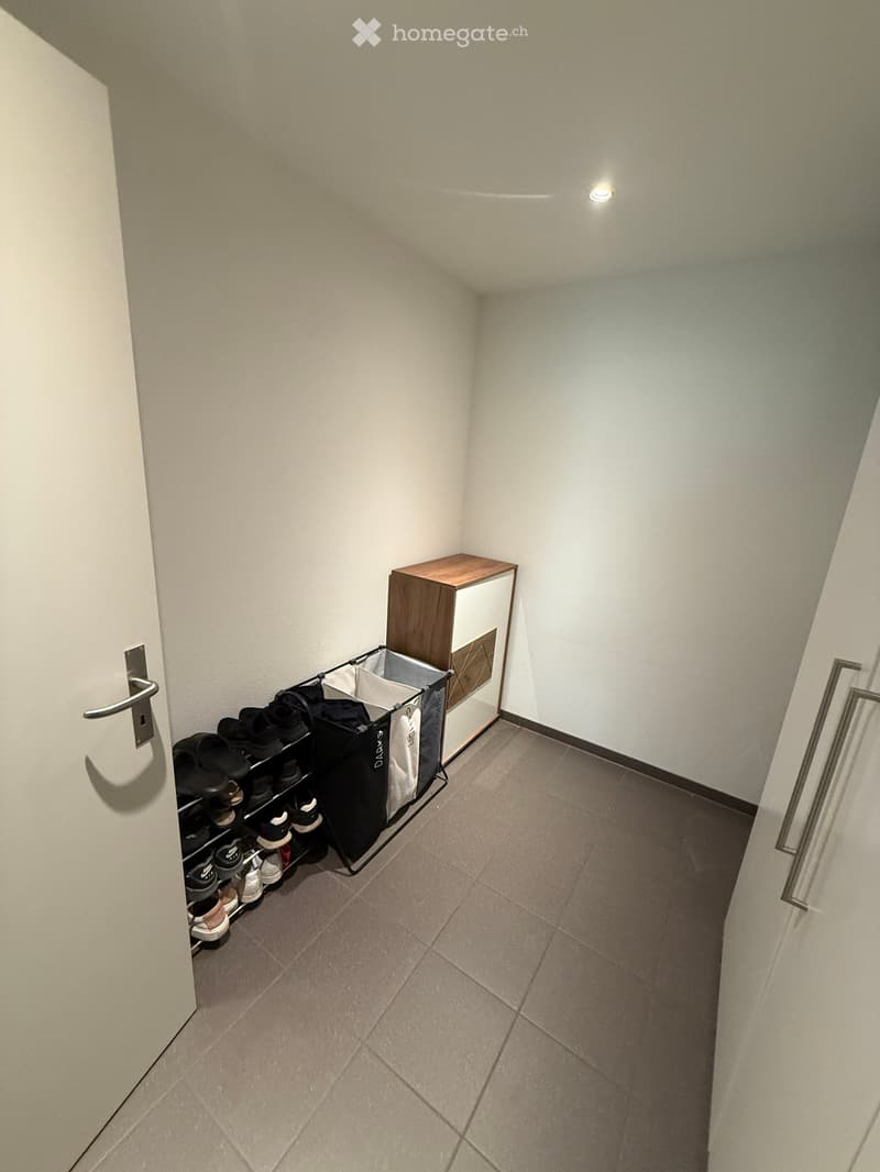 A narrow hallway with tiled flooring, white walls, and a small wooden cabinet with a shoe rack and laundry basket.