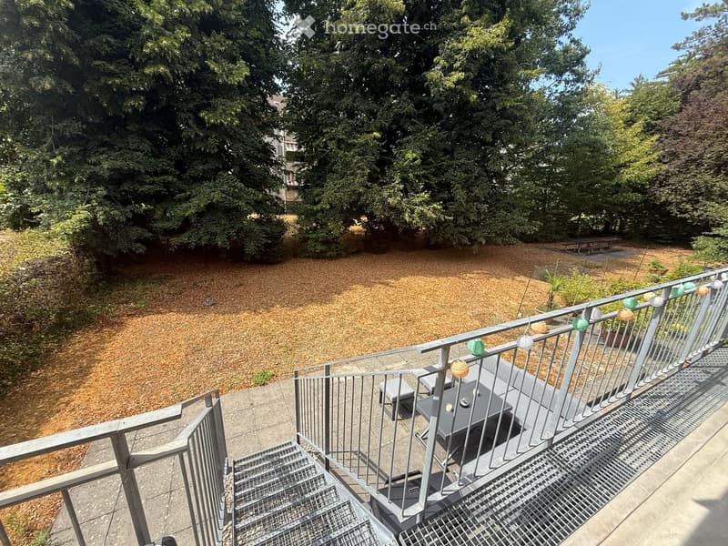 Balcony with a table and chair, overlooking a grassy area with trees and a playground in the background.